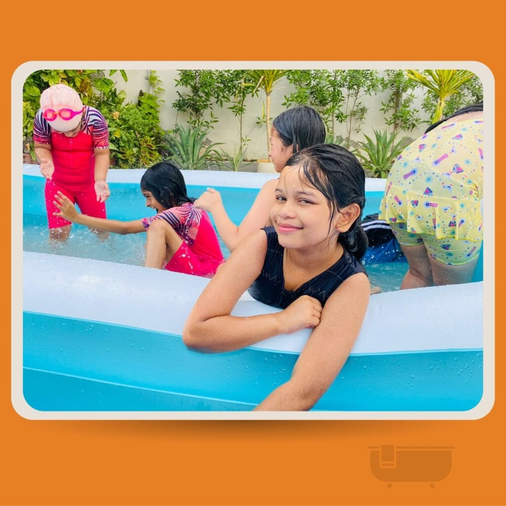 A group of young children having fun and splashing water at each other inside a large blue inflatable swimming pool outdoors.