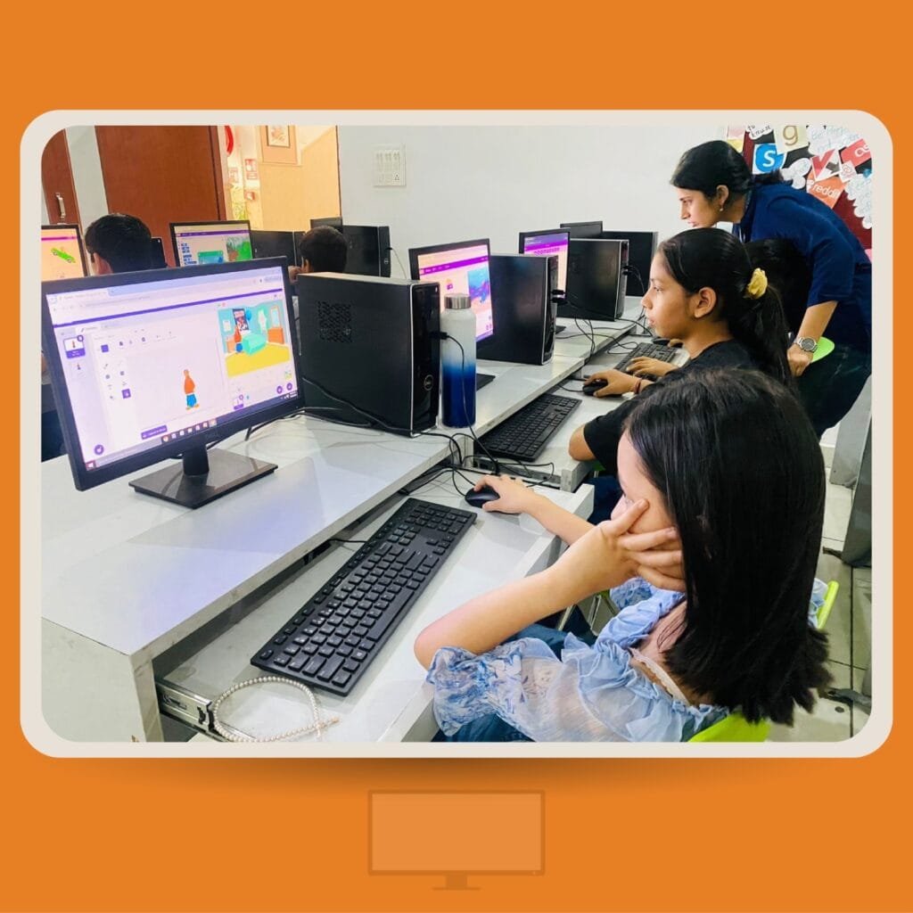 A group of students in a computer lab working on desktop computers. They are using block-based coding software, and a teacher leans in to assist one of the students.
